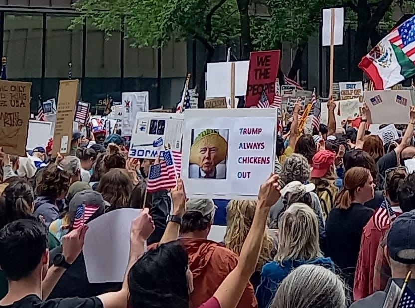 Snapshot of a protest happening in Chicago with a bunch of signs raised. The sign in the middle of th protest reads "Trump Always Chickens Out"